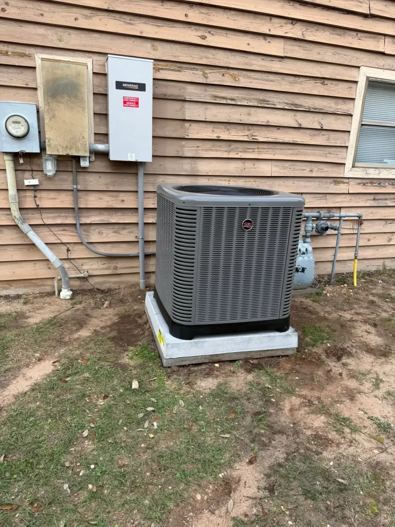 A grey Ruud AC unit and three electrical boxes are installed on a light brown wooden house.